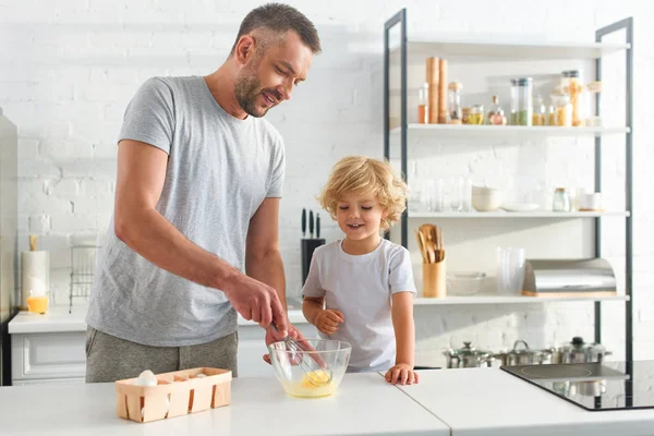 Pai e filho batendo ovos na tigela na mesa na cozinha — Fotografia de Stock