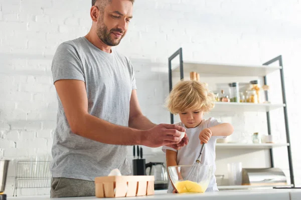 Pequeño niño batiendo huevos mientras padre rompiendo huevo en un tazón en la cocina - foto de stock