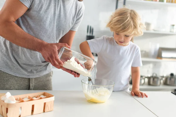 Imagem cortada do homem derramando farinha em tigela enquanto seu filho batendo ovos na cozinha — Fotografia de Stock