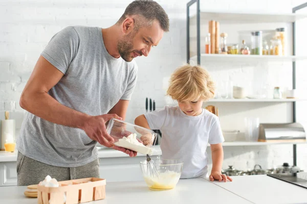 Hombre vertiendo harina en un tazón mientras su hijo batiendo huevos en la cocina - foto de stock