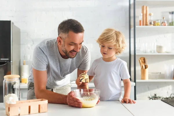 Sonriente hombre sosteniendo tazón mientras su hijo batiendo masa en la cocina - foto de stock