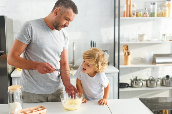 Father holding whisk over bowl while his son trying to lick whisk with dough at kitchen — Stock Photo