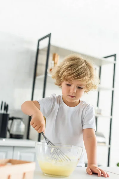 Niño concentrado haciendo masa por batidor en la mesa en la cocina - foto de stock