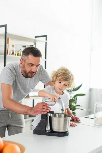 Focused man helping his son using mixer for making dough at kitchen — Stock Photo