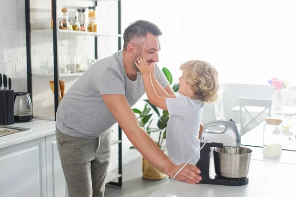 Side view of little boy touching smiling father cheeks near tabletop at kitchen — Stock Photo