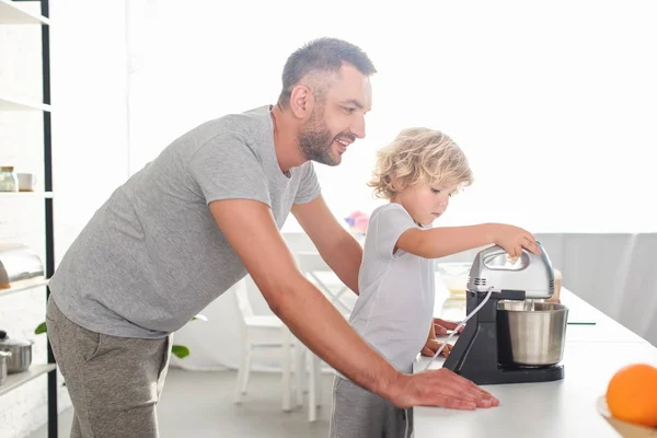 Vista lateral do homem sorridente em pé perto do filho, enquanto ele usa misturador para fazer massa na cozinha — Fotografia de Stock