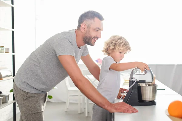 Vue latérale de l'homme souriant debout près de fils alors qu'il utilise mélangeur pour faire de la pâte à la cuisine — Photo de stock