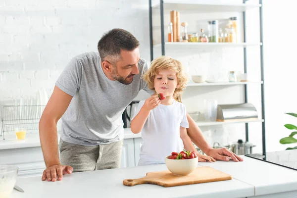 Pai de pé perto do filho enquanto ele come morango na cozinha — Fotografia de Stock