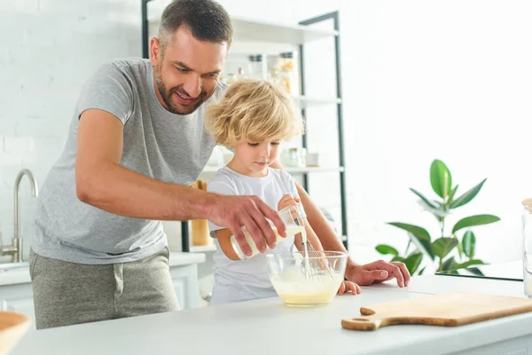 Adorável menino batendo massa enquanto seu pai derramando leite em tigela na cozinha — Fotografia de Stock