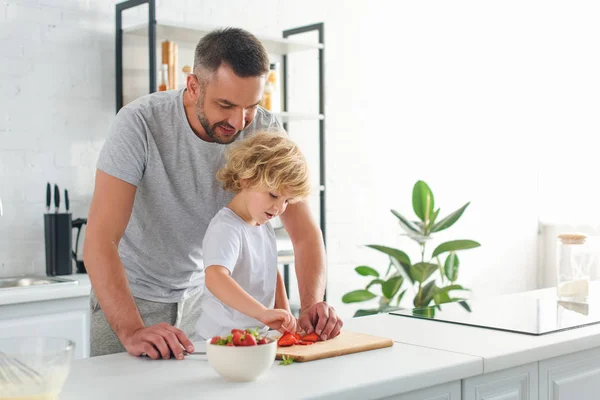 Man standing with son while he taking strawberry from cutting board at kitchen — Stock Photo