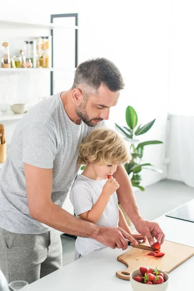 Padre cortando fresa por cuchillo mientras su hijo estaba cerca y comiendo fresa en la cocina - foto de stock