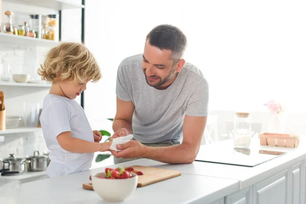 Lächelnder Vater hilft Sohn beim Händewischen mit Serviette neben Tischplatte mit Erdbeeren in der Küche — Stockfoto