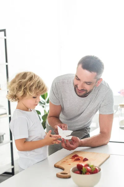 Father helping son to wiping hands by napkin near tabletop with strawberries at kitchen — Stock Photo