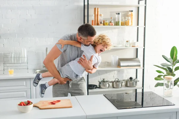 Отец держит смеющегося сына и щекочет его на кухне — стоковое фото