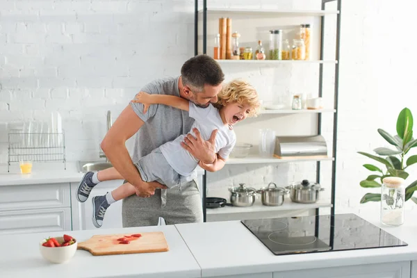 Padre sosteniendo riéndose hijo y cosquillas él en cocina - foto de stock