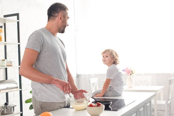 Padre batiendo masa en un tazón y hablando con su hijo mientras está sentado en la mesa en la cocina - foto de stock