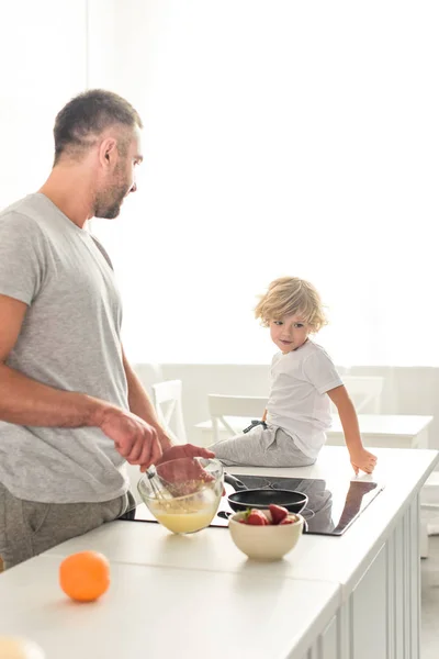 Vista lateral del padre batiendo masa en un tazón y hablando con su hijo mientras está sentado en la mesa en la cocina - foto de stock