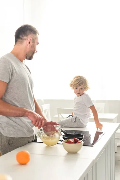 Side view of father whisking dough in bowl and talking to son while he sitting on tabletop at kitchen — Stock Photo