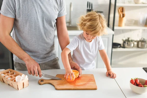 Обрезанное изображение человека с ножом и его сына, берущего кусок апельсина с разделочной доски на кухне — стоковое фото
