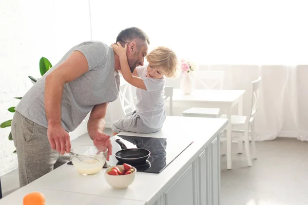 Hijo abrazando sonriente padre mientras él batiendo masa en un tazón en la cocina - foto de stock