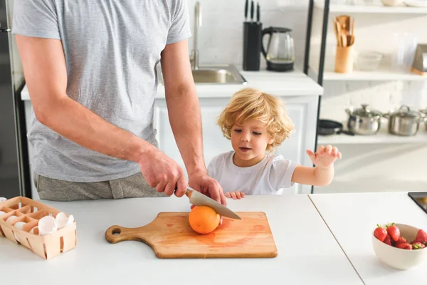Imagen recortada del hombre cortando naranja por el cuchillo y el hijo de pie cerca de la cocina - foto de stock
