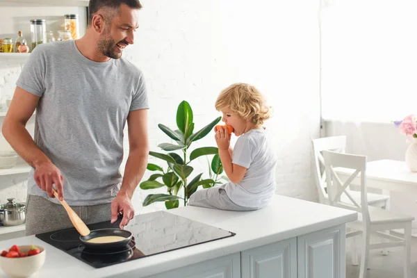 Uomo fare frittelle in padella mentre suo figlio seduto vicino sul tavolo in cucina — Foto stock