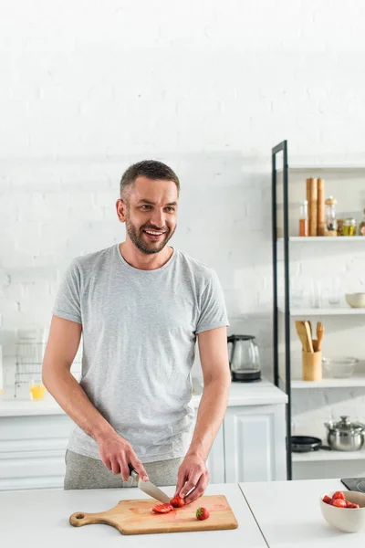 Smiling adult man cutting strawberry by knife on table at kitchen — Stock Photo