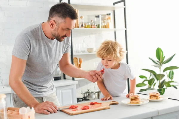 Pai cortando morango e dando peça para filho na cozinha — Fotografia de Stock