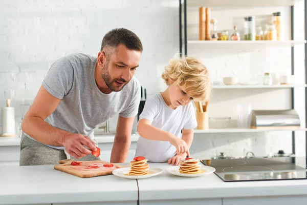 Focused little boy with father putting strawberry pieces on pancakes at kitchen — Stock Photo