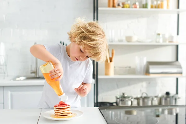 Petit garçon verser du sirop sur des crêpes avec des morceaux de fraise sur l'assiette à la cuisine — Photo de stock