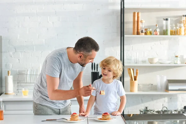 Улыбающийся отец кормит маленького сына блинами на кухне — стоковое фото