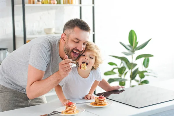 Sonrientes padre e hijo comiendo panqueques cerca de la mesa en la cocina - foto de stock