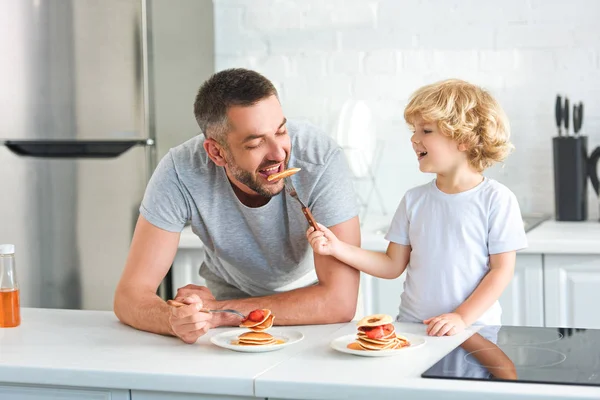 Smiling little boy feeding father by pancake at kitchen — Stock Photo