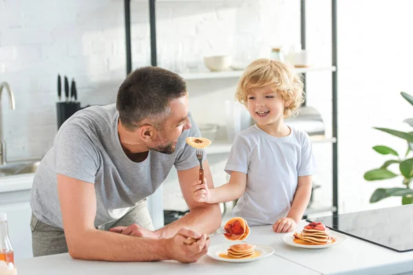 Feliz niño alimentando padre por panqueque en la cocina - foto de stock