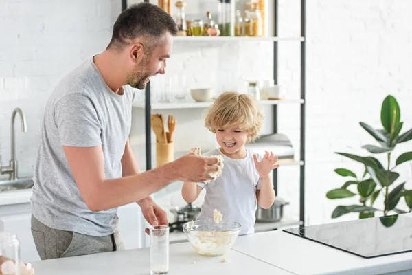 Смеющиеся отец и сын веселятся, делая тесто в миске на кухне — стоковое фото