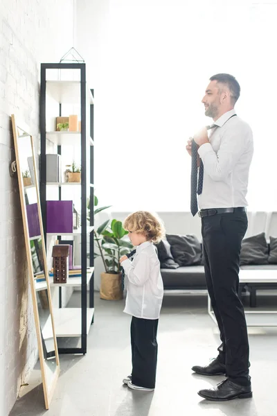 Vista lateral de padre e hijo atando corbatas sobre camisas blancas frente al espejo en casa - foto de stock