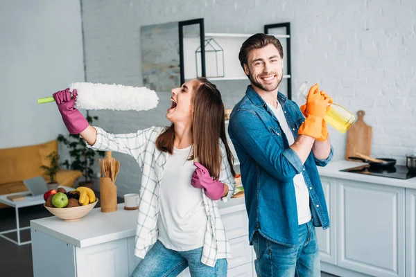 Casal se divertindo durante a limpeza cozinha e namorada fingindo cantar com escova de poeira — Fotografia de Stock