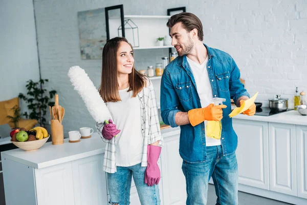 Feliz pareja sosteniendo herramientas de limpieza y mirándose en la cocina - foto de stock