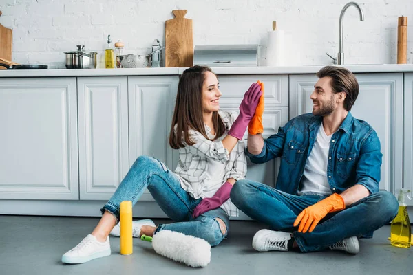 Pareja sentada en el suelo y dando cinco altos después de la limpieza en la cocina - foto de stock