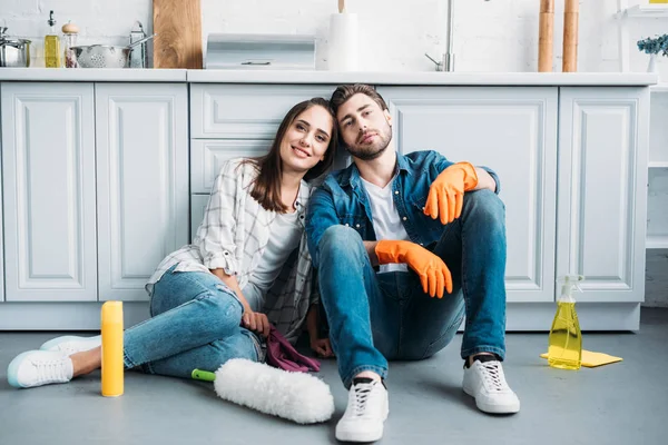 Lächelndes Paar, das nach dem Putzen in der Küche auf dem Boden sitzt und sich auf den Küchentisch lehnt — Stockfoto