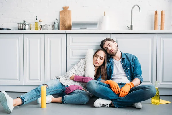 Уставшая пара, сидящая на полу и опирающаяся на кухонный стол после уборки на кухне — стоковое фото