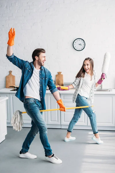 Couple dans des gants en caoutchouc s'amuser avec balai et brosse à poussière dans la cuisine — Photo de stock