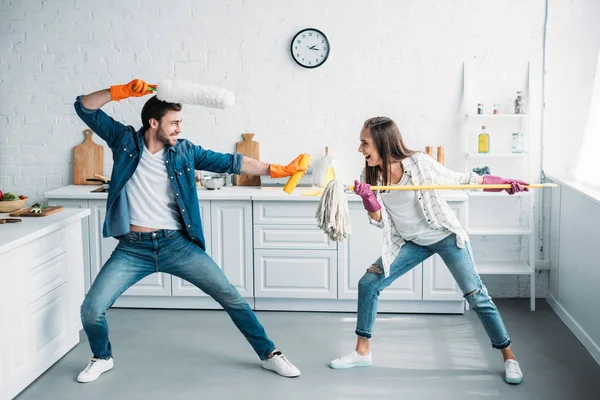 Casal se divertindo e fingindo lutar com ferramentas de limpeza na cozinha — Fotografia de Stock