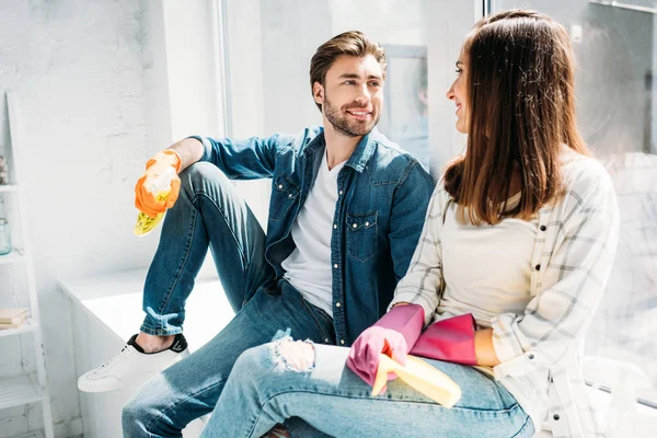 Pareja sentada en el alféizar de la ventana con botella de spray y trapo en la cocina - foto de stock