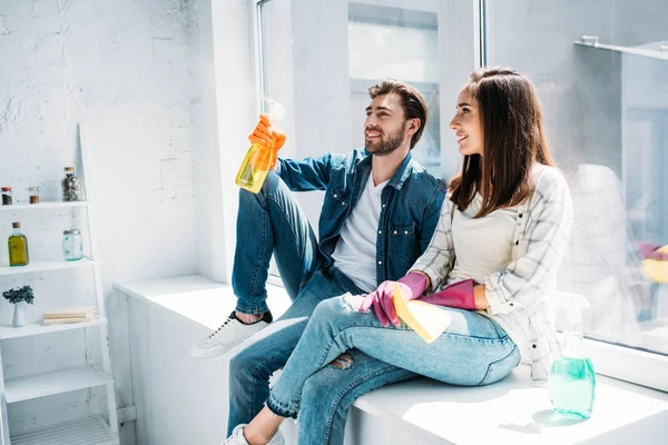 Pareja descansando en el alféizar de la ventana después de la limpieza y divertirse con la botella de aerosol en la cocina - foto de stock