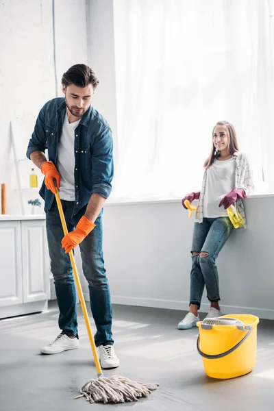 Novio limpieza piso en cocina con fregona y novia apoyada en el mostrador de cocina - foto de stock