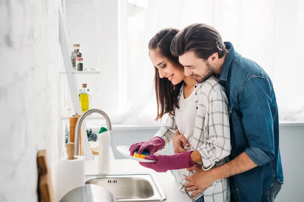 Novia lavando platos y novio abrazándola en la cocina - foto de stock
