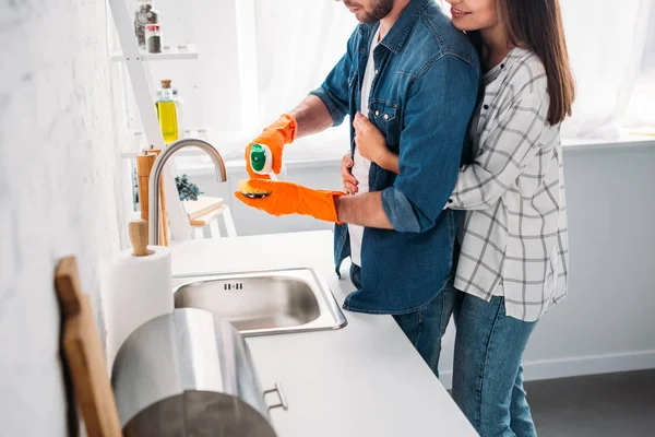 Immagine ritagliata del fidanzato che lava i piatti e la ragazza che lo abbraccia in cucina — Foto stock