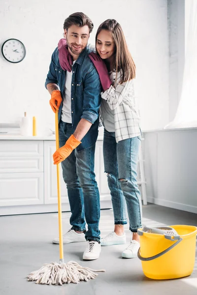 Boyfriend cleaning floor in kitchen with mop and girlfriend hugging him — Stock Photo