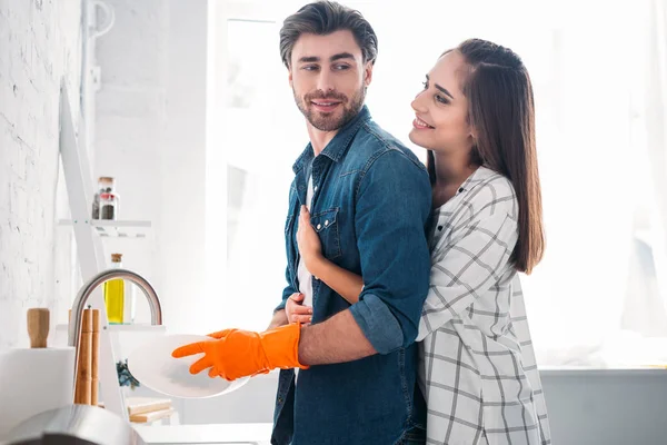 Boyfriend washing dishes and girlfriend hugging him in kitchen — Stock Photo